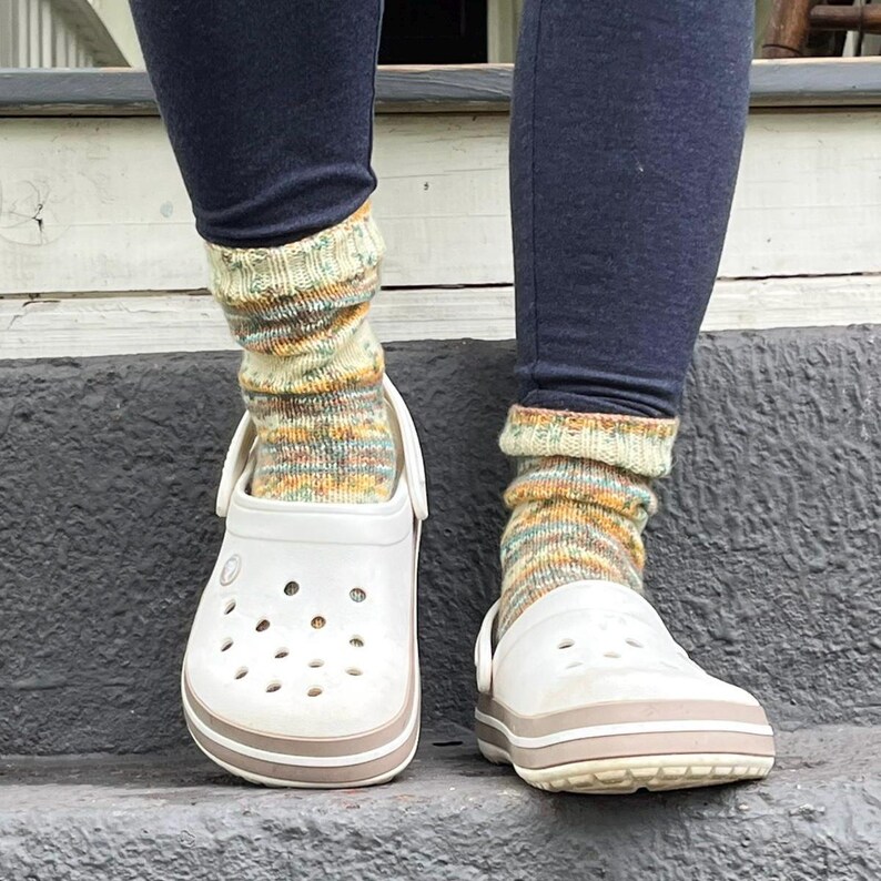 Pu&ograve; includere: Zoccoli bianchi con finiture beige indossati con calzini a maglia colorati. I calzini hanno un mix di tonalit&agrave; gialle, verdi e arancioni. La persona indossa leggings blu scuro. Gli zoccoli sono su una superficie grigia.