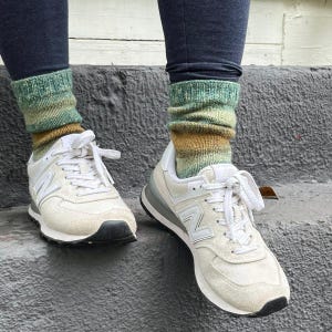 Pu&ograve; includere: Primo piano di scarpe da ginnastica bianche e grigie con calzini verdi, marroni e blu. Le scarpe da ginnastica hanno lacci bianchi e una suola nera. I calzini sono lavorati a maglia con un motivo sfumato.