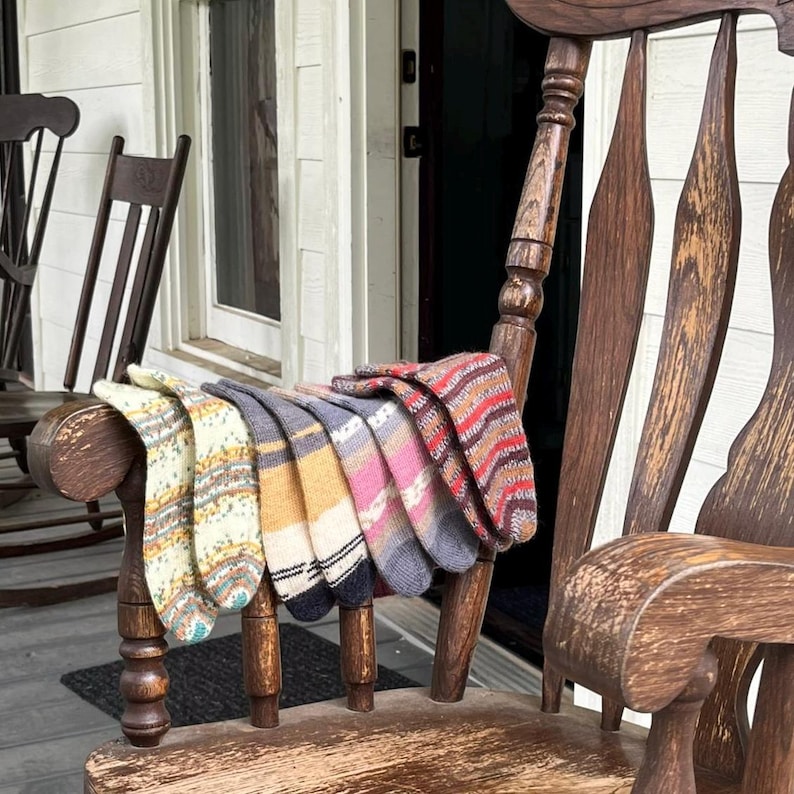 Pu&ograve; includere: Una collezione di calzini colorati e fantasia esposti su una sedia a dondolo in legno. I calzini presentano vari disegni, tra cui righe, motivi geometrici e maglie strutturate. I colori includono crema, giallo, rosa, grigio e rosso.
