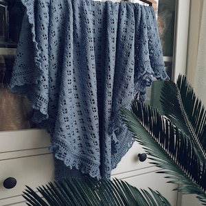 以下が含まれることがあります： 白い背景に、木製のハンガーにかけられた、繊細なレース模様の青いクロシェのショール。ショールは、黒い引き出しの取っ手が付いた白いドレッサーにかけられています。手前に緑色のヤシの葉があります。