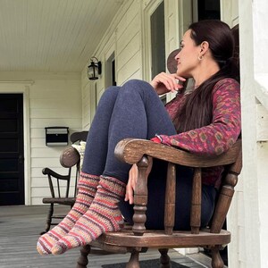 Pu&ograve; includere: Calzini lavorati a mano con strisce orizzontali nei toni del rosso, arancione e beige. I calzini sono indossati con leggings blu scuro. La persona &egrave; seduta su una sedia a dondolo di legno su un portico.