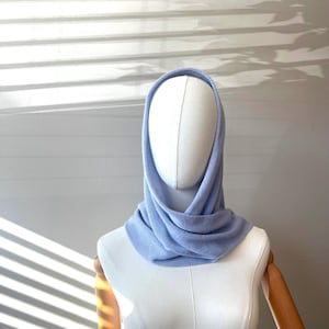 以下が含まれることがあります： マネキンに展示された、水色のニットフードとネックウォーマーの組み合わせ。 ネックウォーマーは首に巻き付けられ、フードは頭を覆っています。 生地は柔らかい質感で、全体的なデザインはシンプルでエレガントです。