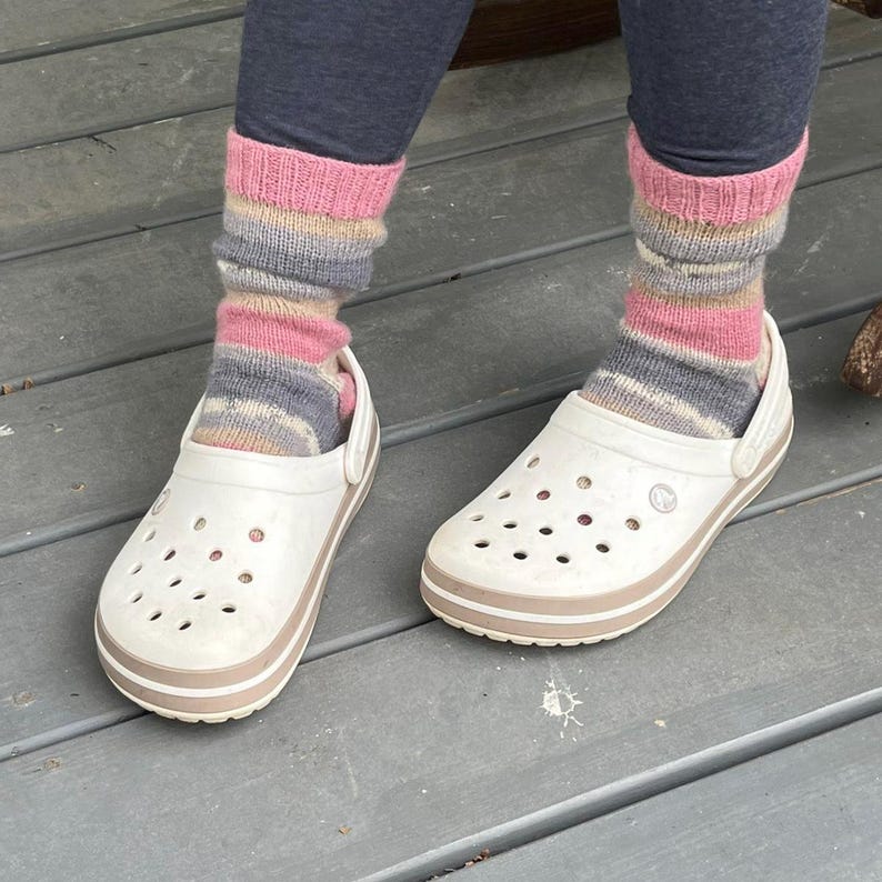 Pu&ograve; includere: Zoccoli bianchi con finiture marroni sono abbinati a scaldamuscoli a righe colorate in rosa, grigio e marrone. Gli zoccoli hanno fori di ventilazione multipli e un cinturino al tallone girevole. Gli scaldamuscoli sono tirati su sopra le caviglie.