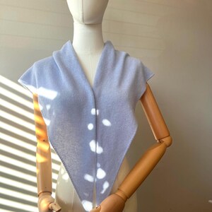 以下が含まれることがあります： Vネックデザインのライトブルーのニットショール。木製の腕を持つマネキンにショールが展示されています。ショールは柔らかいパステルカラーで、軽量素材で作られているようです。ショールは三角形の形をしています。