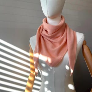 以下が含まれることがあります： マネキンにドレープされたピーチ色の三角形のスカーフ。スカーフは柔らかいニット素材でできており、首の周りにスタイルされています。マネキンは白で、木製の腕が付いています。