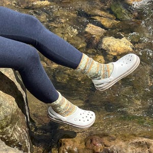 Pu&ograve; includere: Zoccoli bianchi con finiture marroni sono indossati con calzini fantasia. Gli zoccoli hanno un cinturino e sono indossati con leggings blu scuro. La scena &egrave; ambientata vicino a un ruscello.