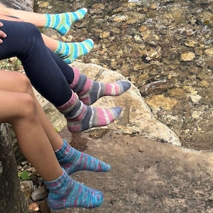 Pu&ograve; includere: Sono visibili diversi paia di calzini colorati a maglia, con strisce e motivi blu, verdi, rosa e grigi. I calzini sono indossati da persone sedute vicino a un ruscello roccioso. I calzini sono un mix di stili.