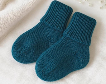 May include: A pair of teal knitted baby socks. The socks feature a ribbed cuff and a soft, textured appearance. They are presented on a white surface, with a glimpse of a white knitted item in the background.