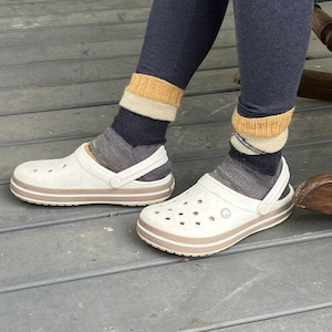 Pu&ograve; includere: Zoccoli bianchi con rifiniture marroni e cinturino, indossati con calzini a righe nei toni del grigio, blu navy e giallo. Gli zoccoli hanno fori multipli e sono su una superficie di legno. Le calze sono tirate su sopra le caviglie.
