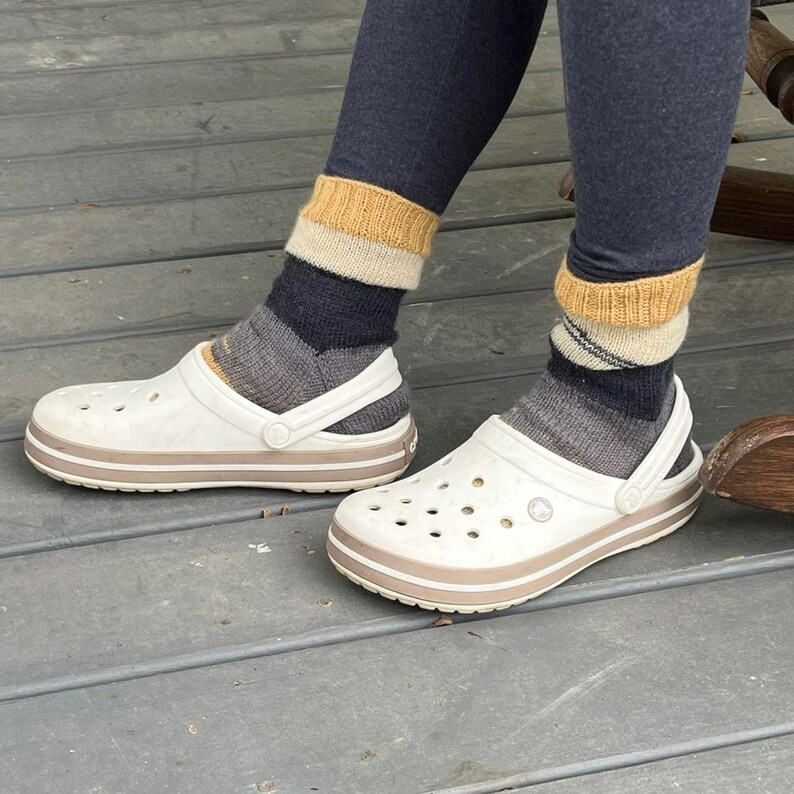 Pu&ograve; includere: Zoccoli bianchi con rifiniture marroni e cinturino, indossati con calzini a righe nei toni del grigio, blu navy e giallo. Gli zoccoli hanno fori multipli e sono su una superficie di legno. Le calze sono tirate su sopra le caviglie.