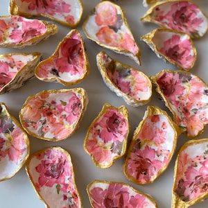 Peut inclure: Plusieurs coquilles d'huîtres décorées d'un motif floral rose et pêche. Les bords de chaque coquille sont peints en or, ajoutant une touche d'élégance. Les coquilles sont disposées sur une surface blanche.