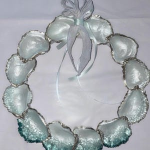 May include: A circular wreath constructed from individual oyster shells with iridescent white interiors and silver-toned edges. The shells are arranged in a ring and adorned at the top with a light blue ribbon tied into a decorative bow.