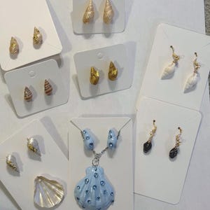 Könnte beinhalten: Eine Sammlung von Schmuck im Muschel-Design, präsentiert auf weißen Karten. Der Schmuck umfasst Ohrstecker, Ohrhänger und eine Halskette mit Anhänger. Die Ohrringe und die Halskette weisen verschiedene Farben auf, darunter Gold, Weiß und Blau, einige sind mit kleinen Kristallen verziert.