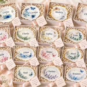 Puede incluir: Una colección de conchas de ostra personalizadas para anillos, cada una en una caja de regalo blanca. Las conchas presentan diseños florales y de mariposas con nombres como "Corina" y "Carolyn". Cada caja incluye una pequeña etiqueta que dice "Thank you for being my Bridesmaid".