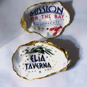 May include: Two white oyster shells with gold-painted edges. One shell has the text "MISSION ON THE BAY SWAMPSCOTT" with a red lobster and stars. The other shell has the text "KLIA TAVERNA" with olive branch art.