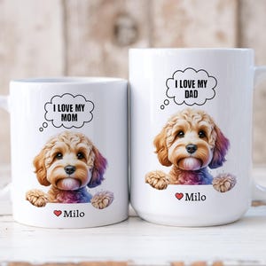 May include: Two white ceramic mugs with a cartoon image of a brown and white dog. The mug on the left says "I love my mom" and the mug on the right says "I love my dad". Both mugs have the name "Milo" printed on the bottom.