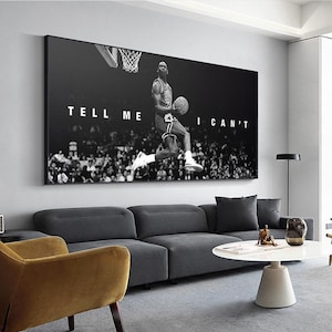 May include: Black and white wall art featuring a basketball player mid-air, about to dunk the ball. The text "TELL ME I CAN'T" is displayed in white block letters. The artwork is displayed above a gray sofa.