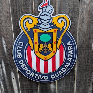 May include: A wooden sign featuring the Club Deportivo Guadalajara logo. The sign has a navy blue border with the words "CLUB DEPORTIVO GUADALAJARA" in white. The center has red and white stripes, a yellow crest, and a green circle.