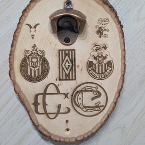May include: A light-coloured oval wood slice wall-mounted bottle opener featuring a bronze-toned metal opener at the top. The wood surface is laser-engraved with various Club Deportivo Guadalajara logos, a goat head, a soccer player, and the letters CD.