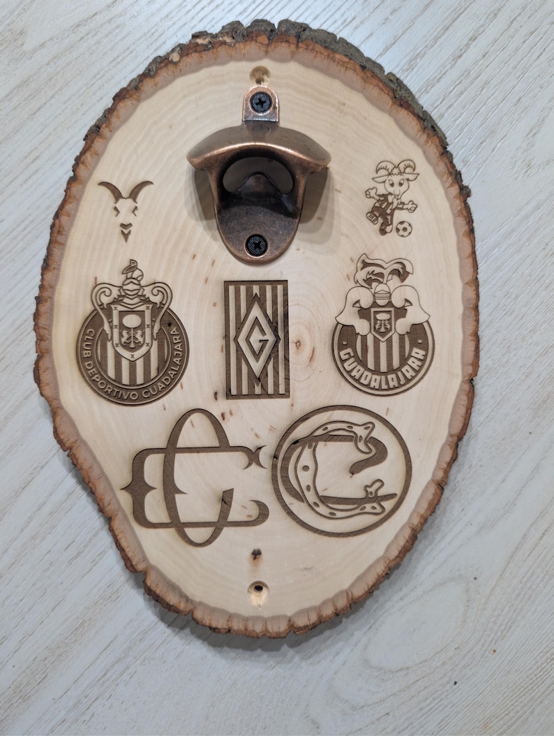 May include: A light-coloured oval wood slice wall-mounted bottle opener featuring a bronze-toned metal opener at the top. The wood surface is laser-engraved with various Club Deportivo Guadalajara logos, a goat head, a soccer player, and the letters CD.