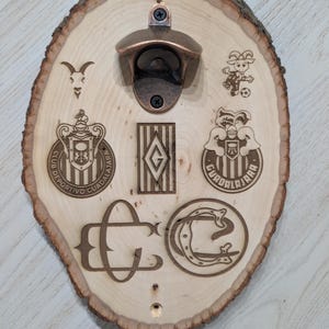 May include: A wall-mounted bottle opener attached to a light-coloured oval wood slice with natural bark edges. The wood features laser-engraved logos, including the Club Deportivo Guadalajara crest, a goat mascot, and various team-related monograms.