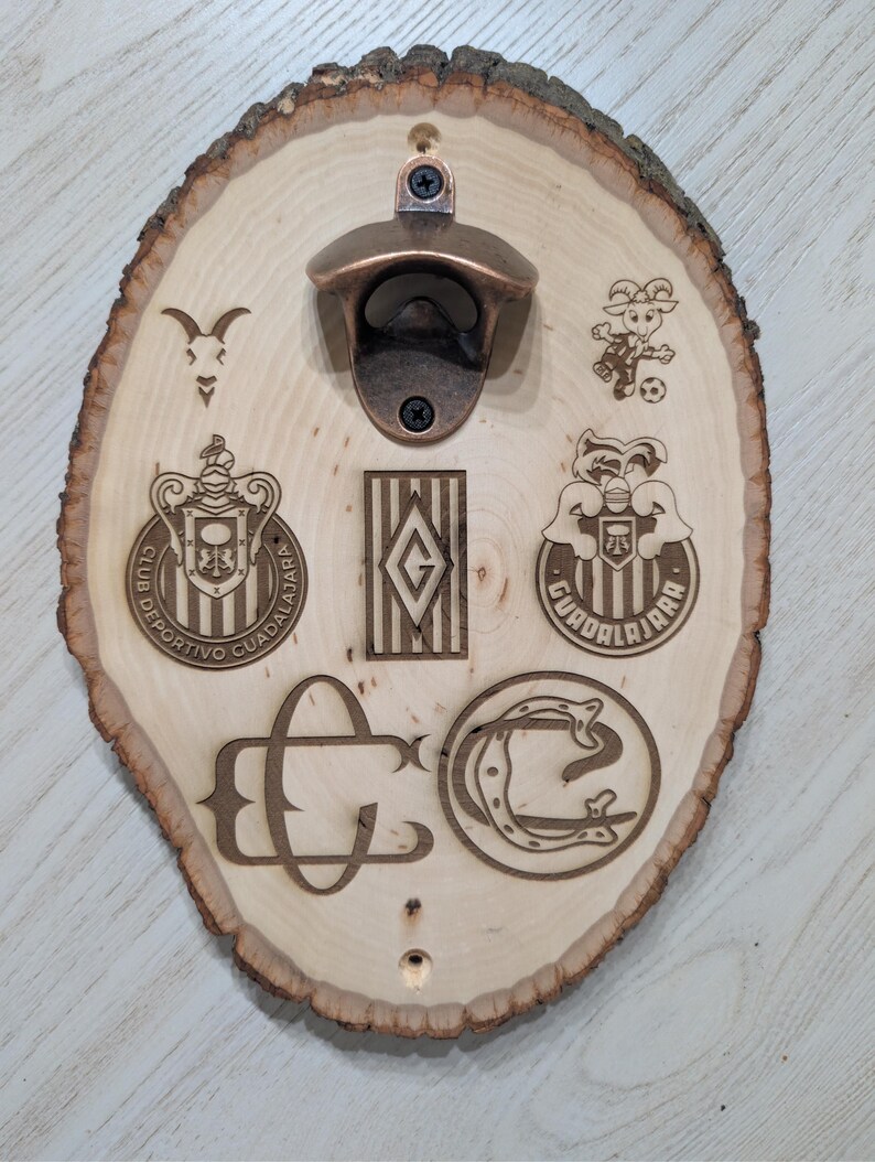 May include: A wall-mounted bottle opener attached to a light-coloured oval wood slice with natural bark edges. The wood features laser-engraved logos, including the Club Deportivo Guadalajara crest, a goat mascot, and various team-related monograms.