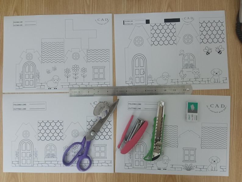 May include: Four white paper sheets with black line art of houses for assembly, featuring a 12-inch metal ruler, purple scissors, a pink stapler, a green utility knife, and a small white eraser. Each sheet includes CAD branding and folding lines.