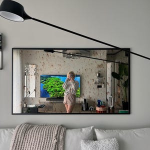 Op de afbeelding: Een zwarte spiegel met lijst die een vrouw weerspiegelt die voor een televisie staat met een afbeelding van een groen landschap. De spiegel hangt aan een witte muur boven een witte bank met een lichtgrijze deken en twee witte kussens.