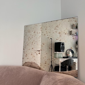 Pode incluir: Um espelho grande e retangular com uma superfície marrom salpicada reflete uma sala com uma caixa de som preta, um suporte de microfone e uma mesa com uma cadeira. O espelho está montado em uma parede branca.