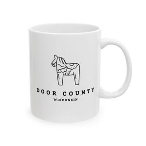May include: White ceramic mug featuring a black line drawing of a Dala horse, with the text "DOOR COUNTY WISCONSIN" in black. The mug has a curved handle and a cylindrical shape.