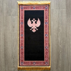 Puede incluir: Una alfombra de oración rectangular con un centro negro sólido y un emblema de águila bicéfala de color rosa claro. La alfombra está enmarcada por un borde decorativo en tonos rosa, azul y blanco, con flecos dorados en ambos extremos.