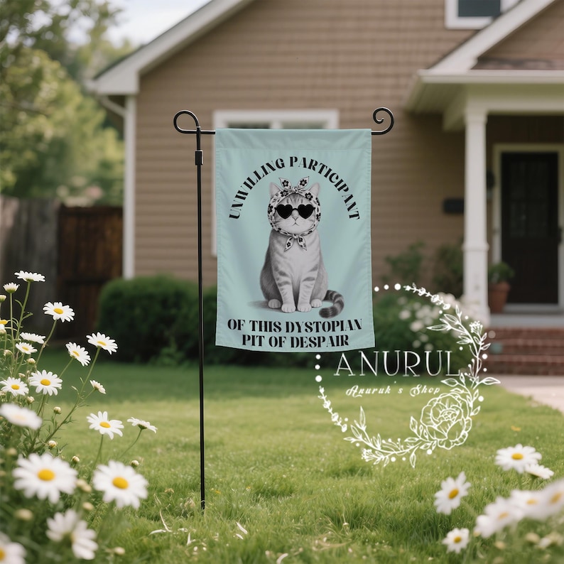 K&ouml;nnte beinhalten: Eine hellblaue Gartenflagge mit einer grauen Katze, die eine herzf&ouml;rmige Sonnenbrille und ein Bandana tr&auml;gt. Auf der Flagge steht "UNWILLING PARTICIPANT OF THIS DYSTOPIAN PIT OF DESPAIR". Die Flagge wird in einem Garten mit G&auml;nsebl&uuml;mchen gezeigt.