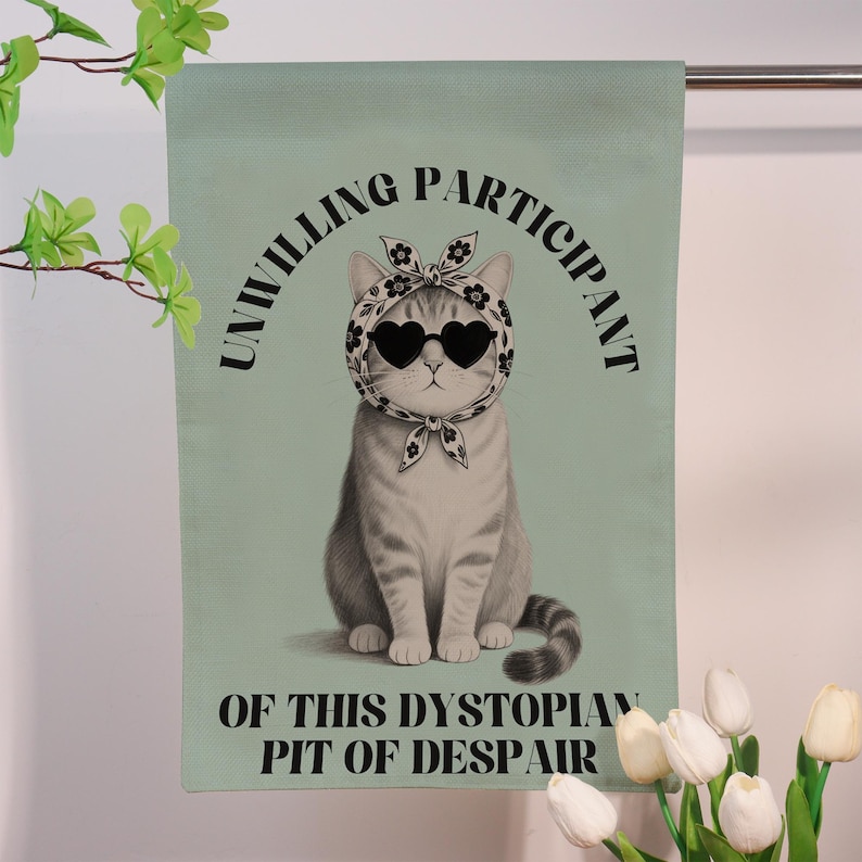 K&ouml;nnte beinhalten: Eine hellgr&uuml;ne dekorative Flagge mit einer grauen Katze, die eine herzf&ouml;rmige Sonnenbrille und ein Kopftuch tr&auml;gt. Der Text lautet "UNWILLING PARTICIPANT OF THIS DYSTOPIAN PIT OF DESPAIR".