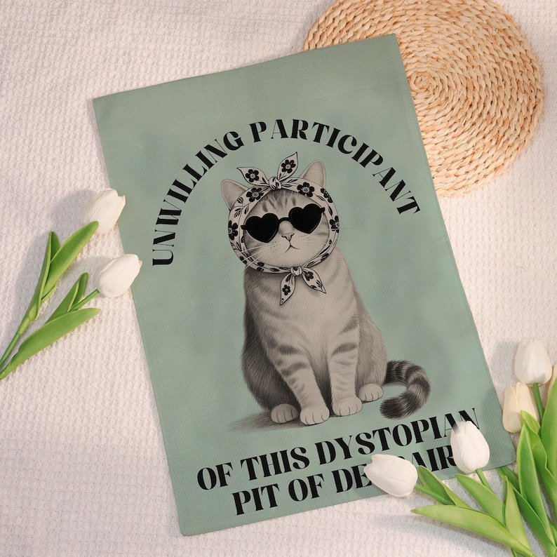 K&ouml;nnte beinhalten: Eine hellgr&uuml;ne rechteckige Flagge mit einer grauen Katze, die eine herzf&ouml;rmige Sonnenbrille und ein Blumenschal tr&auml;gt. Der Text "UNWILLING PARTICIPANT OF THIS DYSTOPIAN PIT OF DESPAIR" ist in Schwarz gedruckt.