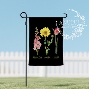 May include: Black garden flag featuring watercolor illustrations of a foxglove, daisy, and tulip. The flowers are in shades of pink, yellow, and red, with green stems and leaves. The word "FOXGLOVE", "DAISY", and "TULIP" are printed in white.