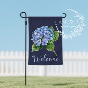 May include: A navy blue garden flag features a watercolor illustration of a blue hydrangea flower with green leaves. The word "Welcome" is written in white script at the bottom. The flag is displayed on a black metal stand.