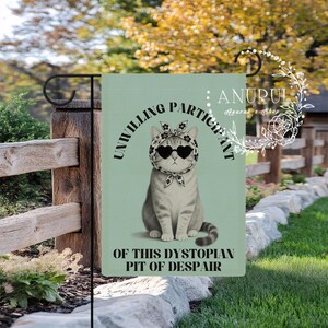 K&ouml;nnte beinhalten: Eine Gartenflagge mit hellgr&uuml;nem Hintergrund zeigt eine Katze mit herzf&ouml;rmiger Sonnenbrille und Schal. Der Text lautet &bdquo;UNWILLING PARTICIPANT OF THIS DYSTOPIAN PIT OF DESPAIR&ldquo;. Die Flagge wird im Freien gezeigt.