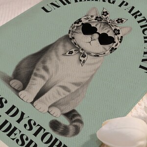 K&ouml;nnte beinhalten: Vintage-Illustration einer grauen getigerten Katze mit herzf&ouml;rmiger Sonnenbrille und einem Blumenschal auf hellblauem Hintergrund. Der Text um die Katze lautet "UNWILLING PARTICIPANT" und "IS DYSTOPIAN DESPAIR".