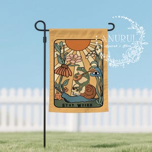 Peut inclure: Un drapeau de jardin jaune moutarde avec une illustration fantaisiste. Une grenouille joue d'un instrument à cordes sur un escargot. Le drapeau porte l'inscription "STAY WEIRD" et un motif floral et solaire.