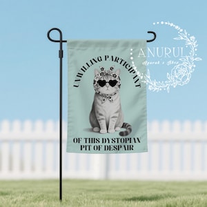 K&ouml;nnte beinhalten: Eine Gartenflagge mit einer grau-wei&szlig;en Katze, die eine herzf&ouml;rmige Sonnenbrille und ein Kopftuch tr&auml;gt. Die Flagge tr&auml;gt den schwarzen Text "UNWILLING PARTICIPANT OF THIS DYSTOPIAN PIT OF DESPAIR". Die Flagge ist an einer schwarzen Metallstange befestigt.