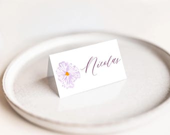 May include: A white place card with a watercolor-style purple flower and the name "Nicolas" in cursive script. The card is folded and rests on a white plate, suitable for weddings or events.