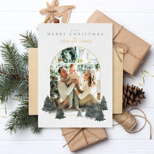 Może przedstawiać: Kartka świąteczna ze zdjęciem rodzinnym w złotym łukowatym obramowaniu. Na kartce widnieje napis "2025 Merry Christmas the Anderson Family". Akwarelowe drzewka iglaste znajdują się na dole. Kartka otoczona jest zapakowanymi prezentami, igłami sosnowymi i szyszką.