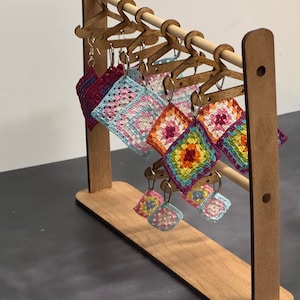 Op de afbeelding: Een houten displaystandaard met gehaakte vierkante oorbellen in verschillende kleuren, waaronder roze, blauw, groen en geel. De oorbellen hangen aan kleine houten hangers.
