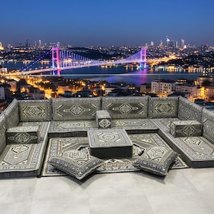 Puede incluir: Un sofá seccional gris y beige con cojines a juego y una mesa de centro, ubicado en una azotea con vistas a una ciudad al anochecer. El sofá y la mesa tienen un diseño estampado. El horizonte de la ciudad incluye un puente iluminado.