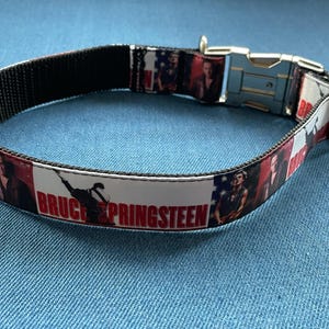 May include: A black and white dog collar with a red and white patterned fabric featuring a picture of Bruce Springsteen and the text "Bruce Springsteen".