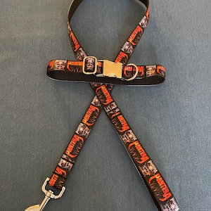 May include: A black and red dog collar and leash set with the word "Pantera" repeated on the fabric. The collar has a silver metal buckle and the leash has a silver metal clip.