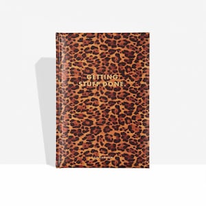 Puede incluir: Un cuaderno de tapa dura con estampado de leopardo. El texto dorado en la portada dice "GETTING STUFF DONE". El cuaderno es de la marca "& Other Stories" y está sobre un fondo blanco.