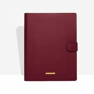 Könnte beinhalten: Ein weinrotes, ledergebundenes Notizbuch mit Magnetverschluss. Das Cover hat eine glatte Textur und eine kleine goldfarbene rechteckige Platte am unteren Rand. Das Notizbuch ist geschlossen und scheint neu zu sein.