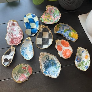 May include: A collection of decorative oyster shells with gold-painted edges. Each shell features a unique design, including floral patterns, checkered patterns, and citrus fruit slices. The shells are arranged on a dark surface.