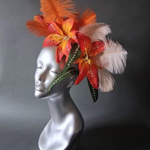 May include: A decorative headpiece featuring orange and red lily flowers, white and orange feathers, and green leaves. The headpiece is adorned with rhinestones and is displayed on a silver mannequin head. The design is suitable for special events.